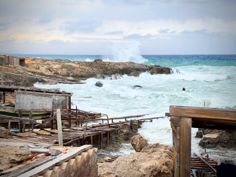 Waves breaking over rocks beside weathered wooden boat sheds and walkways at Es Caló, Formentera